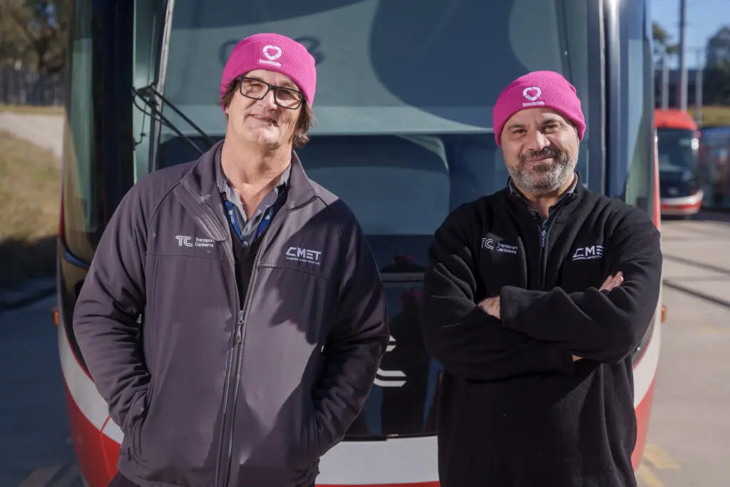 Two CMET team members standing in front of a light rail vehicle, wearing CMET uniform jackets and bright pink beanies. The pair are positioned side by side on a sunny day, with the front of the light rail vehicle visible behind them and tracks extending into the background.