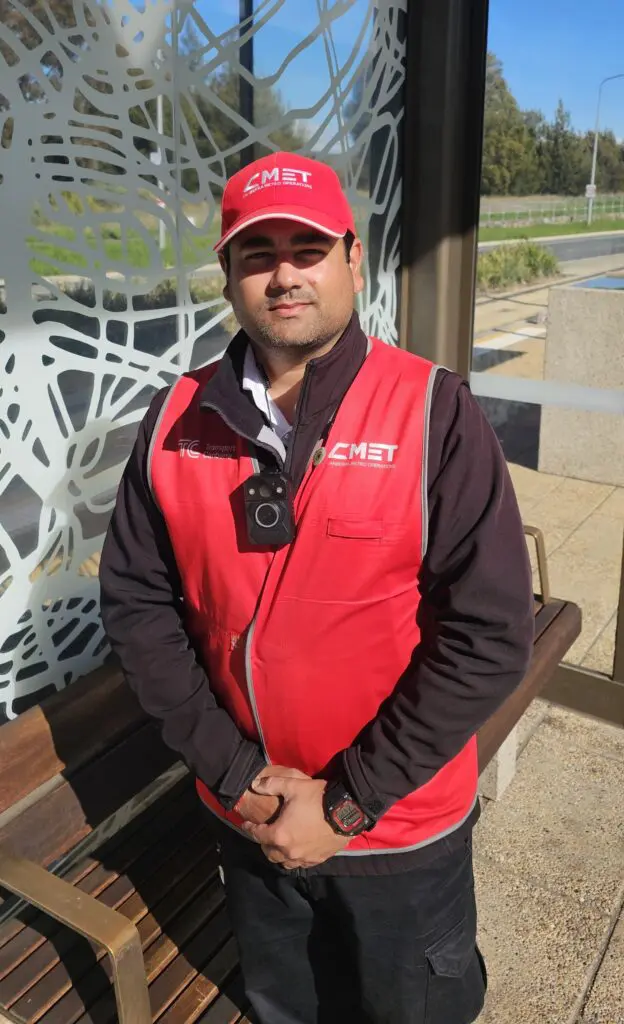 A person wearing a red CMET cap and red CMET safety vest over a dark jacket stands at a bus stop, with a body-worn camera attached to the front of the vest. The person is standing with hands clasped in front of a bench beside a decorative metal screen. A road, footpath and trees are visible in the background on a sunny day.